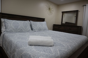 A neatly made bed with a patterned bedspread and pillows, a white towel, a wall clock, and a mirror on a dresser.