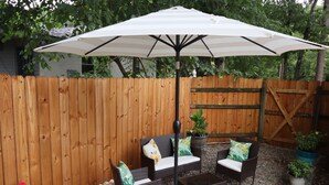 Een terras met houten omheining, twee rieten stoelen met kussens, een grote parasol en potplanten.
