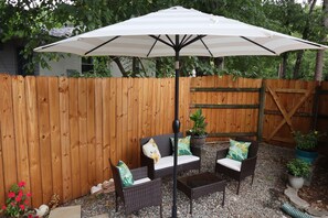 Eine Terrasse mit Holzzaun, zwei Korbstühle mit Kissen, einem großen Sonnenschirm und Topfpflanzen.