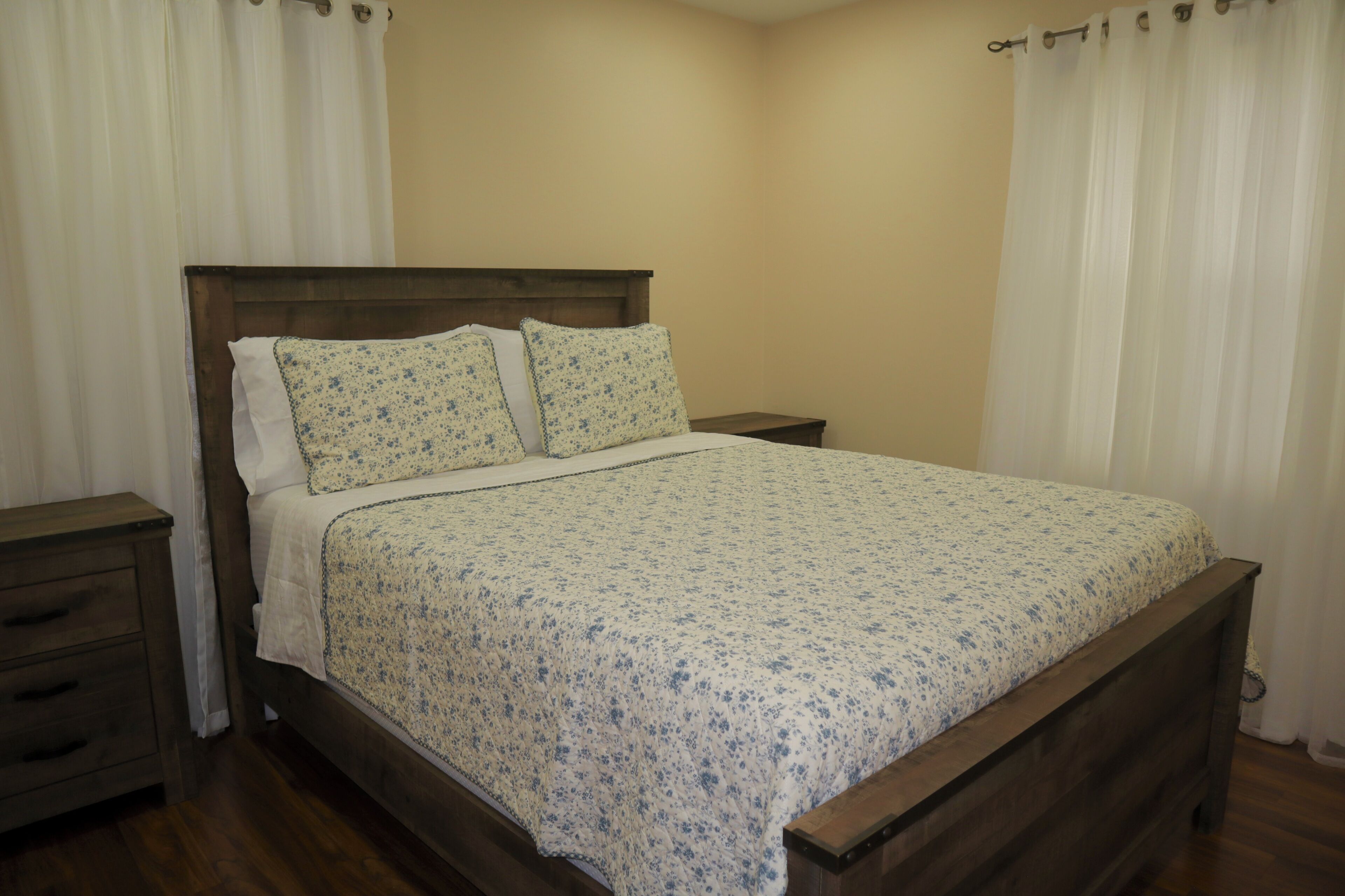 A bedroom with a wooden bed, white curtains, and a wooden nightstand with drawers.
