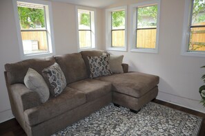A sectional sofa with patterned cushions, a patterned rug, and a view of a wooden fence through windows. - Plan a stay at Westchester Cottage (Charlotte)
