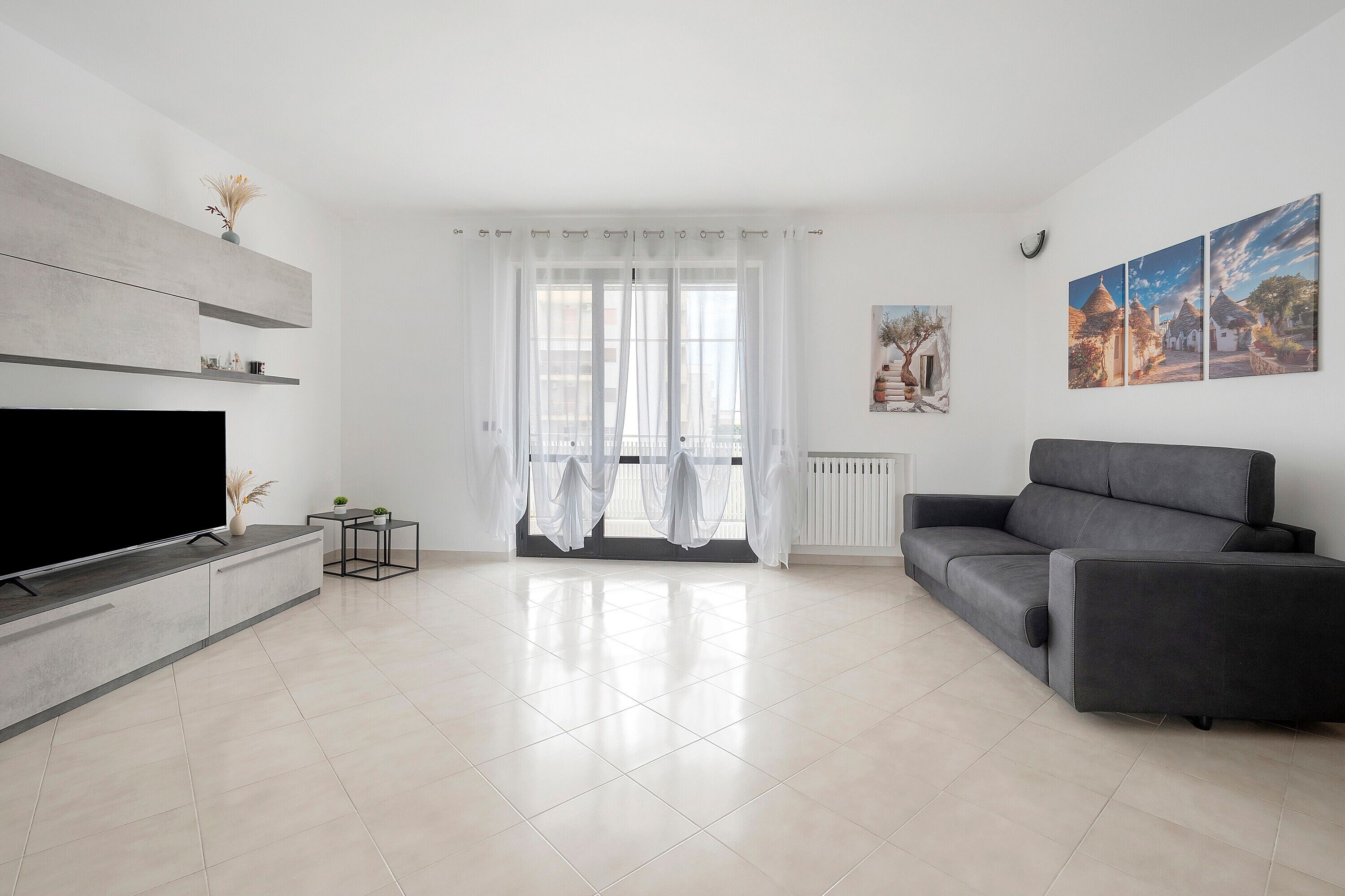 A modern living room with a flat-screen TV, a grey sofa, a coffee table, and a large window with sheer curtains.