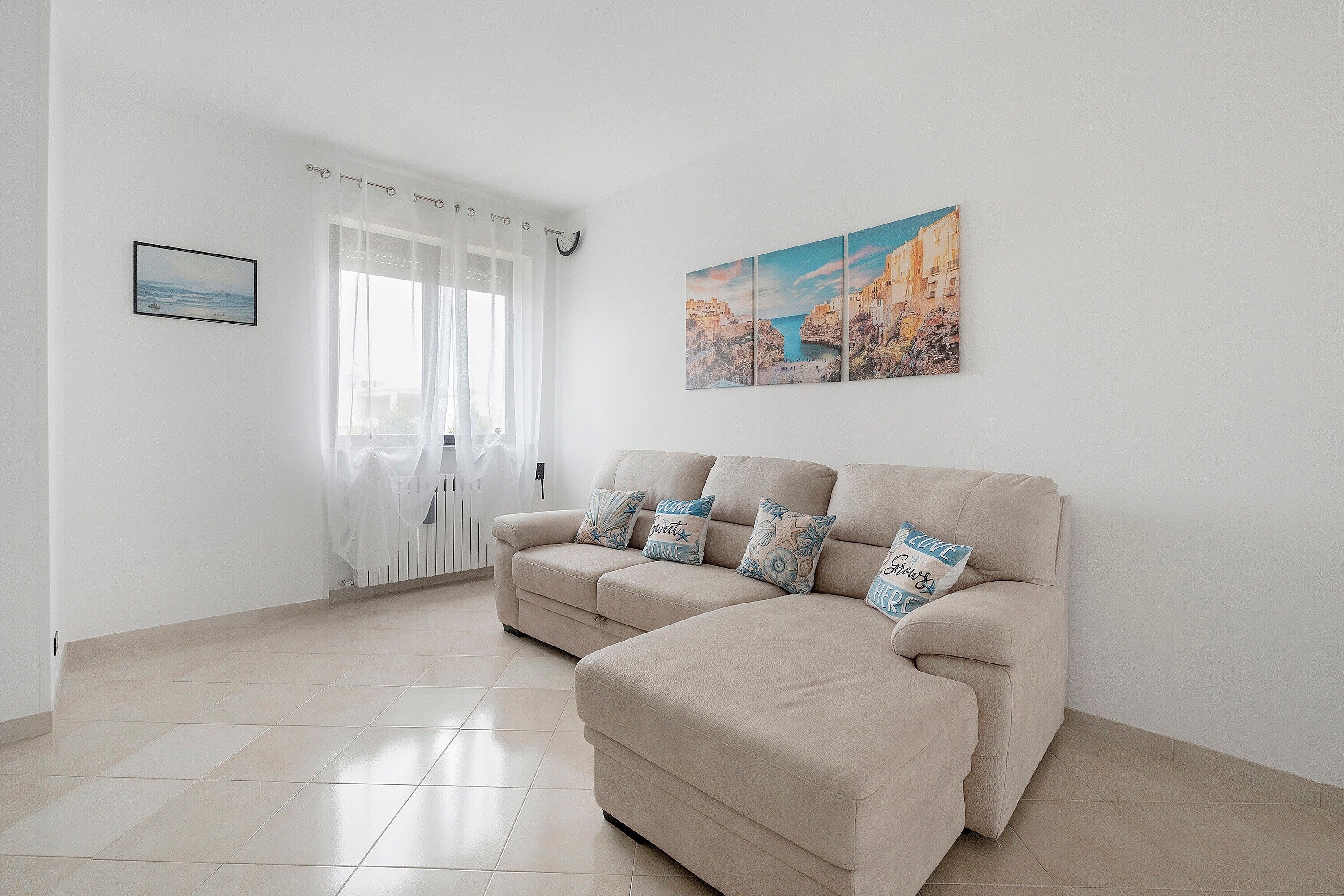 Ein Wohnzimmer mit einem beigen Sofa, einem gerahmten Bild an der Wand und einem Fenster mit transparenten Vorhängen.