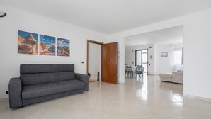 En rummelig opholdsstue med en grå sofa, et spisebord med stole, og et køkken i baggrunden.