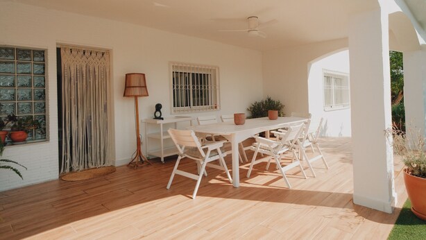 Una habitación amplia con suelo de madera, una mesa blanca con sillas, un ventilador de techo y ventanas con barrotes blancos.