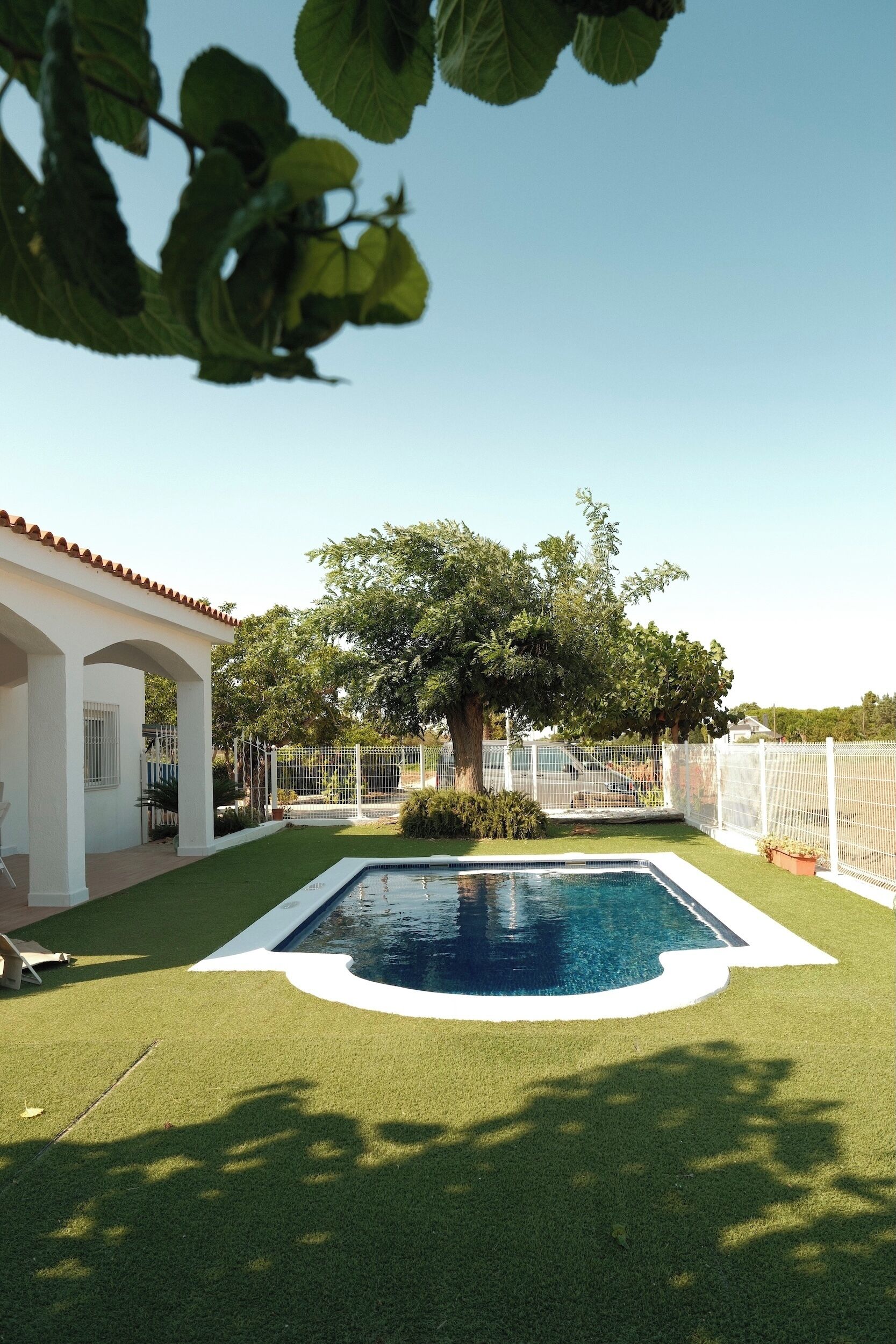 Una piscina rodeada por un césped cuidado, un edificio blanco con techo de tejas y una zona vallada con árboles.
