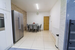 A compact kitchen with a refrigerator, a dining table with chairs, and a wooden door. - Amazing 6 Bed House
Sleeps 14 | Private Parking | EV Charger | Long Stays (London)