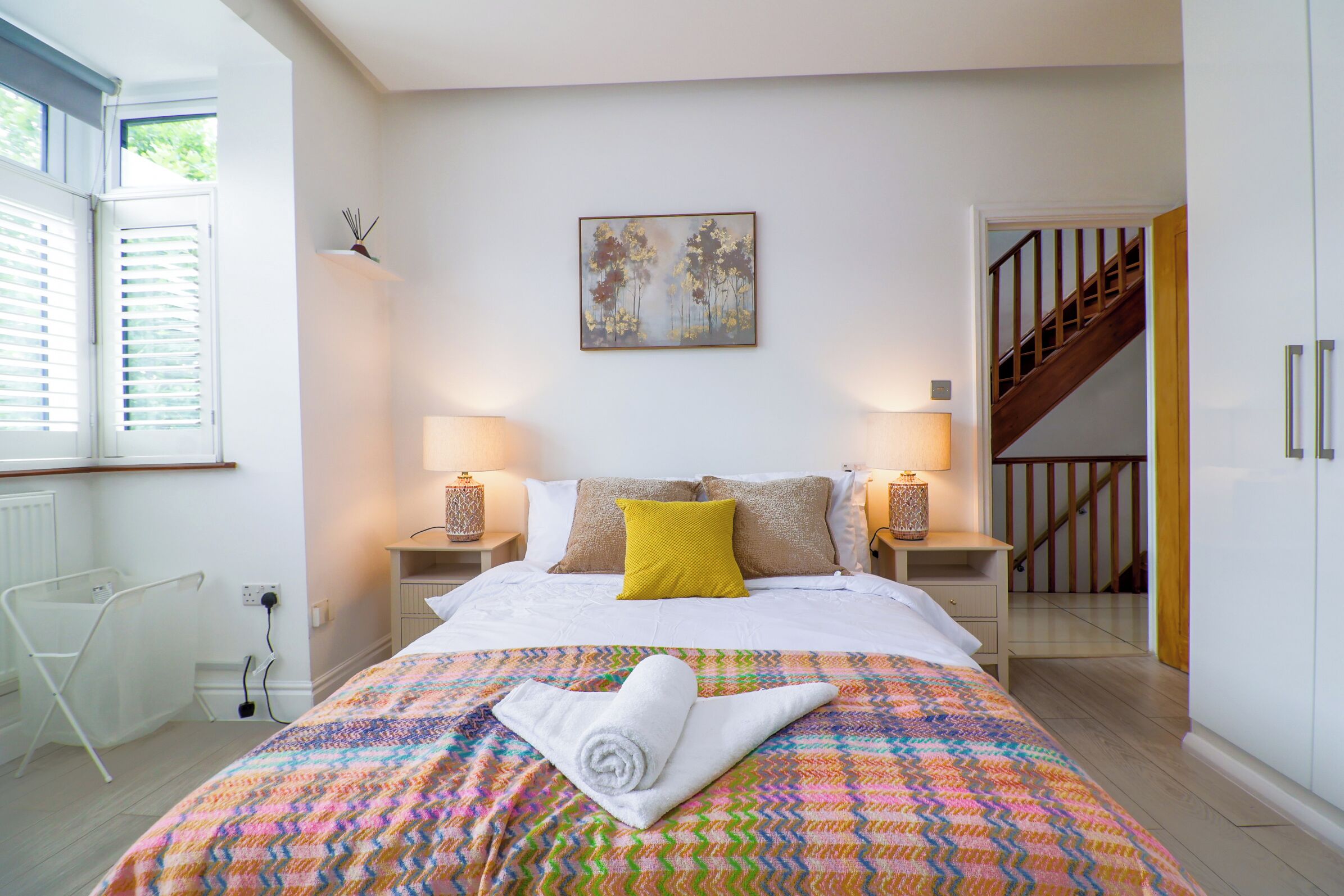Ein Schlafzimmer mit einem Bett, zwei Nachttischlampen, einem Fenster mit Jalousien, einem Bild an der Wand und einer Treppe, die in einen anderen Raum führt.