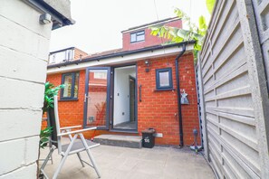 A red brick house with a sliding door, a small garden area with a metal table and a trash bin, and a white garage door. - Amazing 6 Bed House
Sleeps 14 | Private Parking | EV Charger | Long Stays (London)