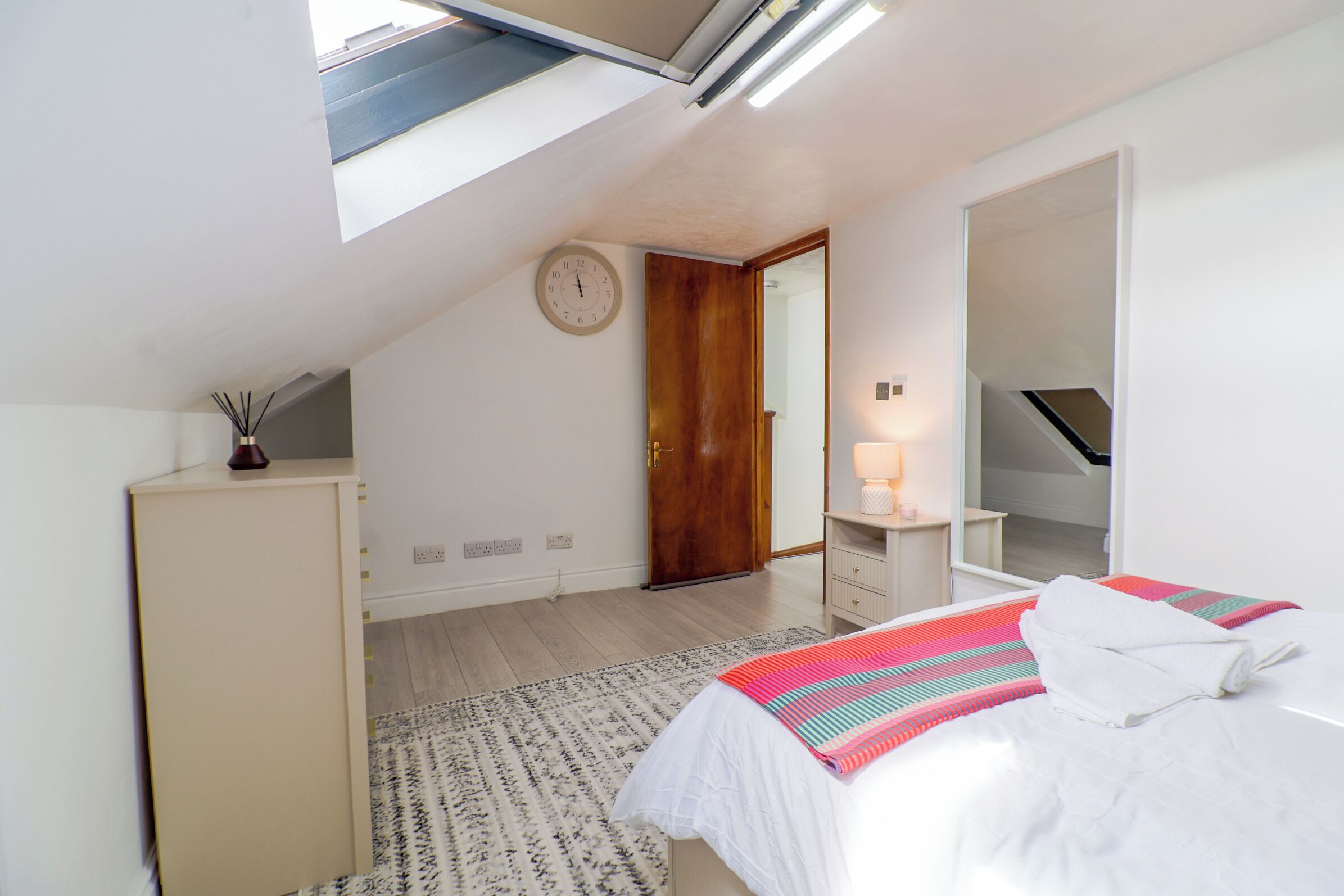A bedroom with a skylight, a bed with a colorful blanket, a wooden door, and a small bedside table with a lamp.