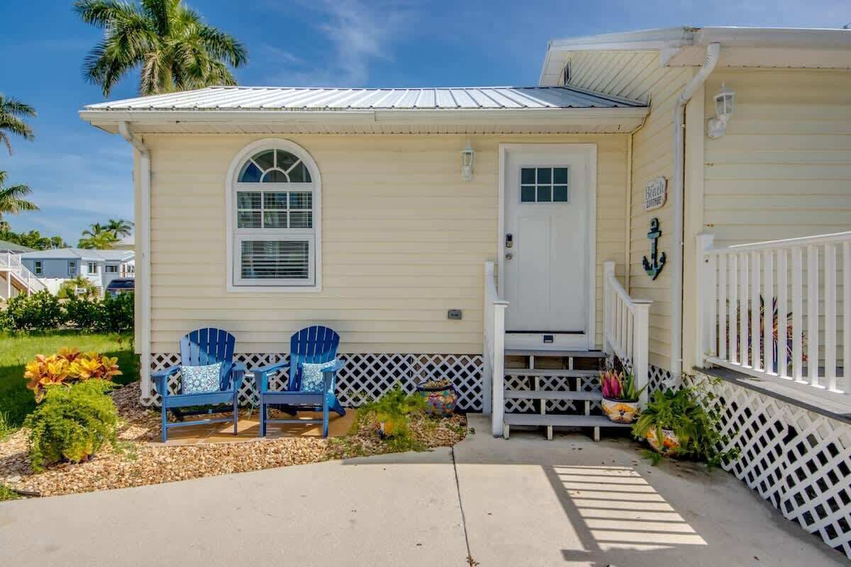 Una casa a un piano con un portico, due poltrone Adirondack, un piccolo giardino e una porta bianca.