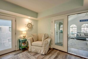 Une pièce avec un fauteuil beige, une petite table avec une lampe, une horloge au mur et une porte-fenêtre coulissante donnant sur un balcon.