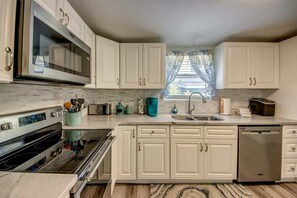 A kitchen with white cabinets, a stainless steel oven, a dishwasher, and a window with curtains. - 214 Pearl Main Pet Friendly Cottage near Beach (Fort Myers Beach)