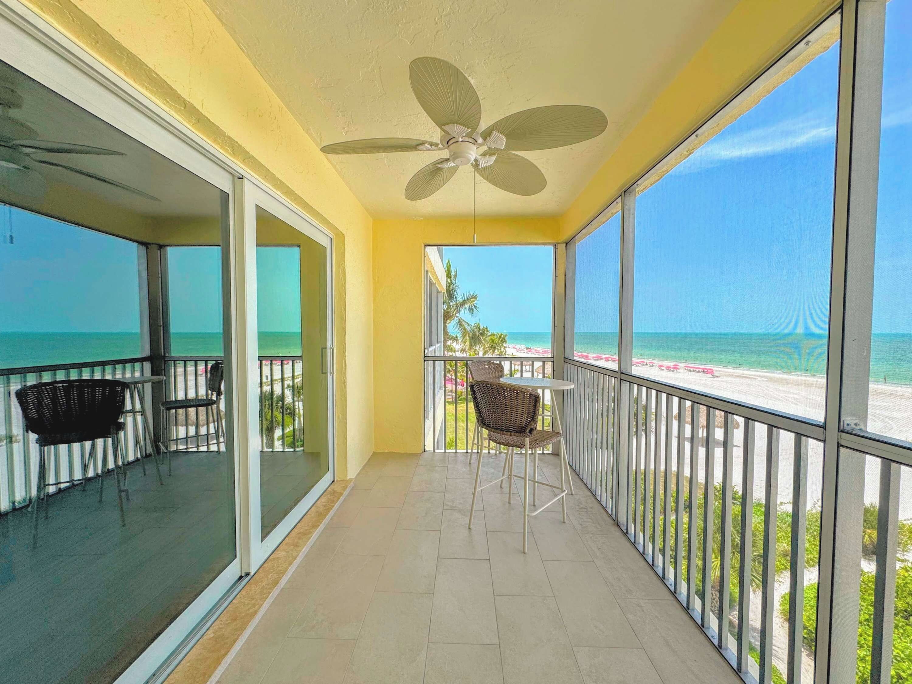 A balcony with a ceiling fan, a table, and chairs, overlooking a beach with clear skies.