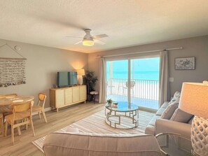 A living room with a TV, a ceiling fan, a dining area with chairs and a table, and a view of the ocean through large sliding doors. - Vacation Villas #432 - Beachfront Condo (Fort Myers Beach)