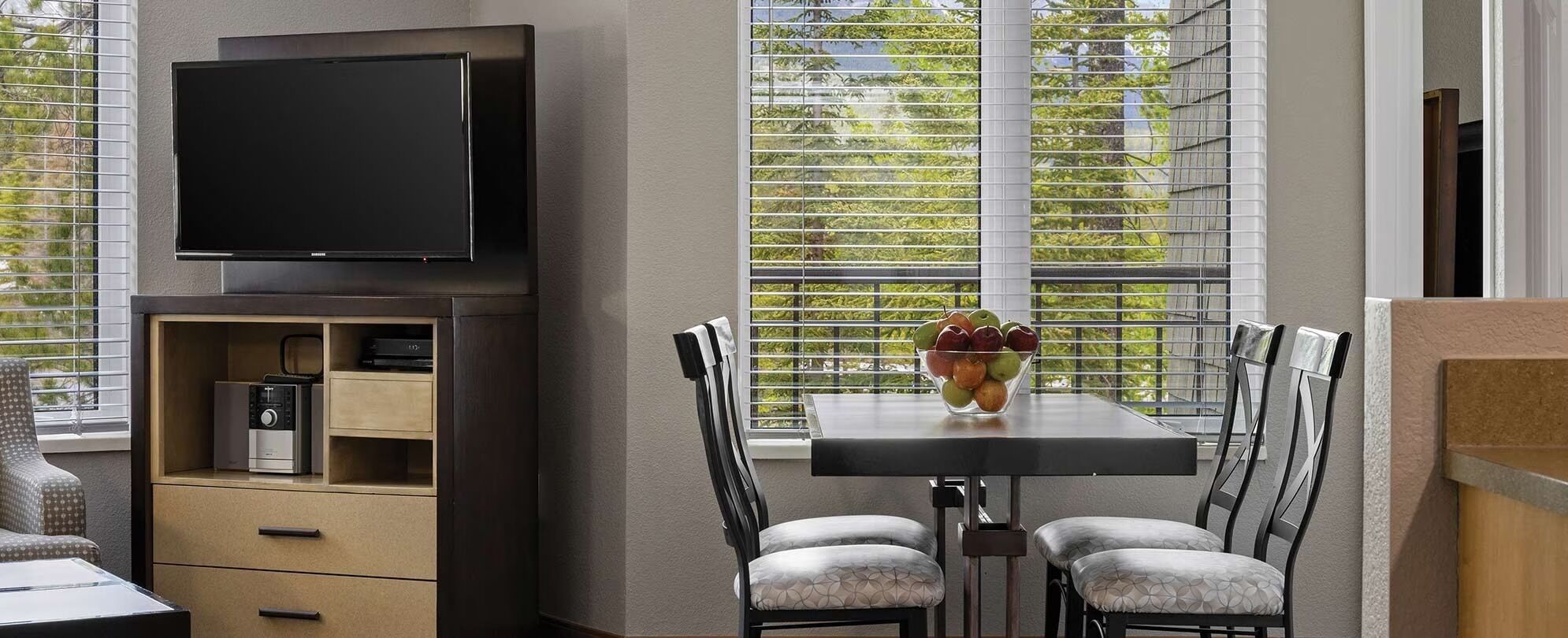 Ein Zimmer mit Flachbildfernseher an der Wand, einem Tisch mit Stühlen und einem Fenster mit Jalousien.
