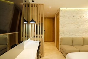 Ein modernes Hotelzimmer mit einem weißen Bett, einem beigen Sofa, einem Fernseher und einem hölzernen Raumteiler mit Pendelleuchten.
