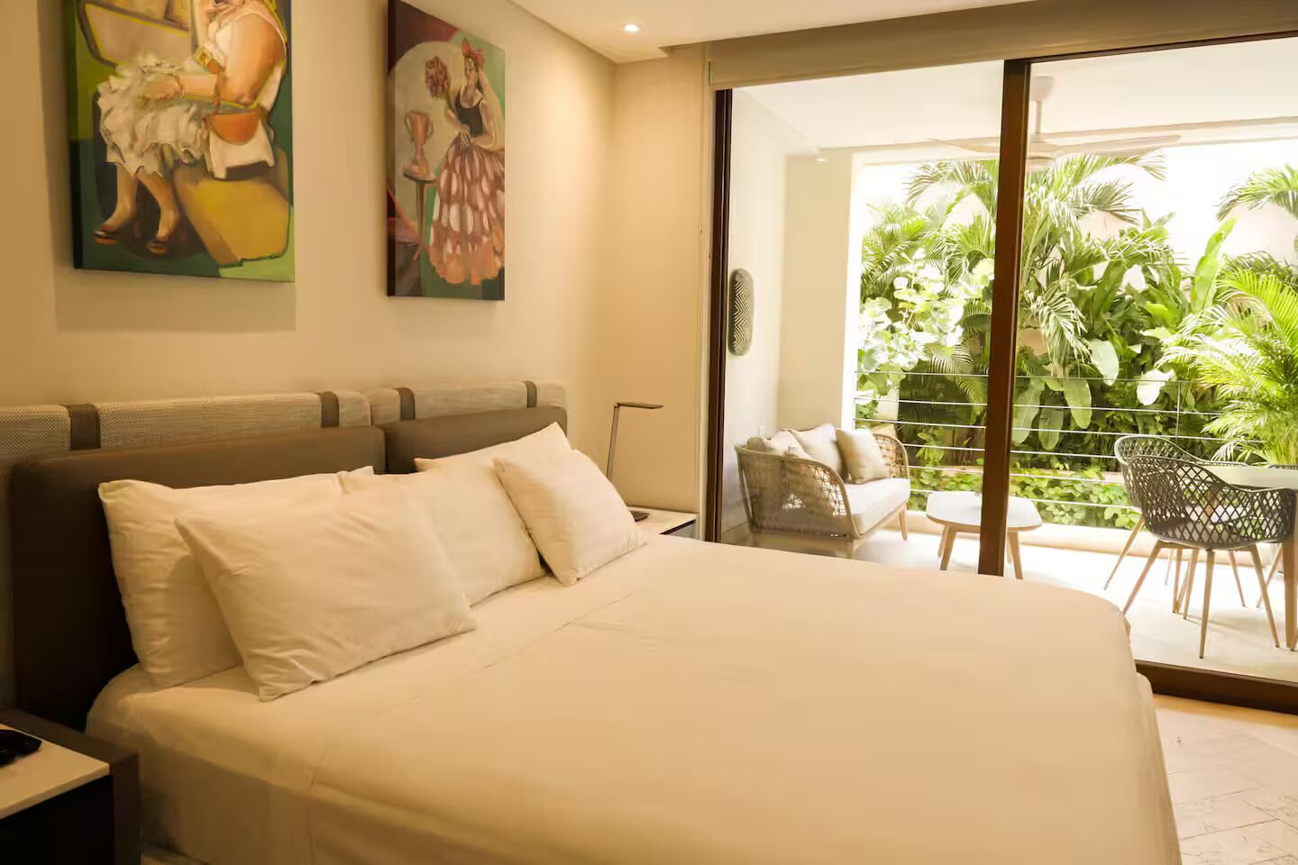 A hotel room with a large bed, a balcony with chairs, and a view of greenery.