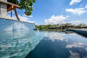 Ein klares Schwimmbecken mit einer gefliesten Kante, einer Holzterrasse und einem Baum im Hintergrund.
