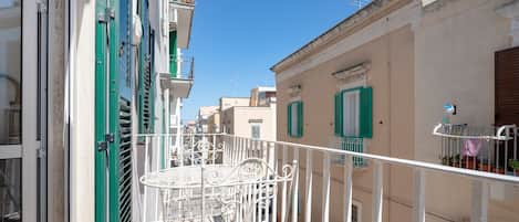 Una calle estrecha flanqueada por edificios, con un balcón que tiene una mesa y sillas de metal blanco, y un cielo azul despejado.