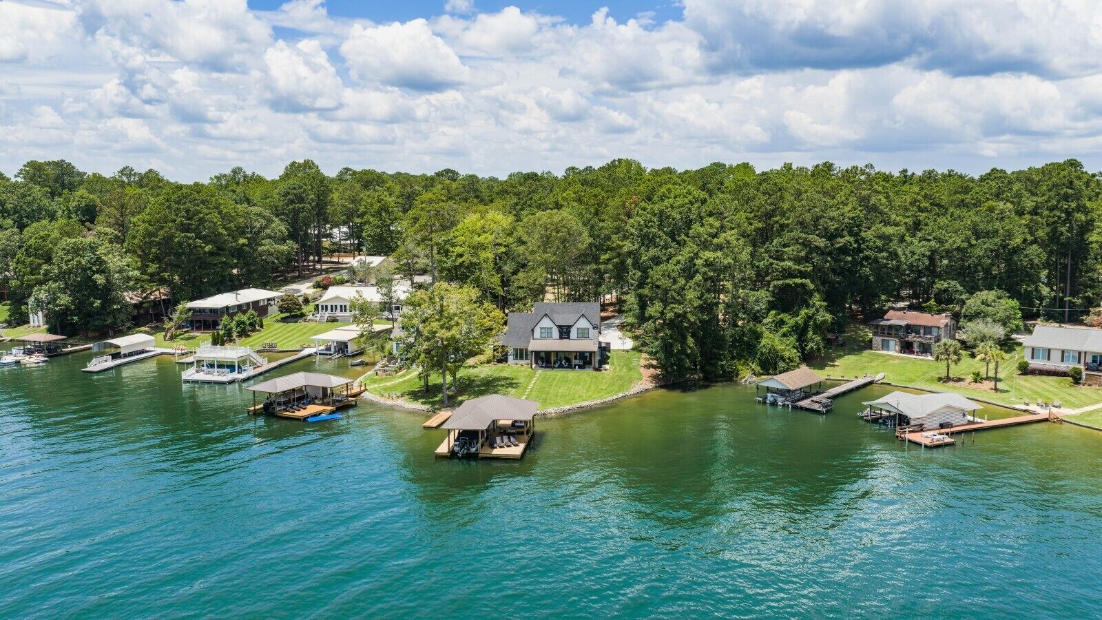 A lakeside property with multiple docked boats, houses, and a dense forest in the background.