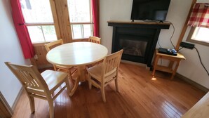 Une pièce comprenant une table ronde en bois, quatre chaises, un téléviseur posé sur un meuble au-dessus d’une cheminée, et une petite table d’appoint en bois équipée d’une multiprise.