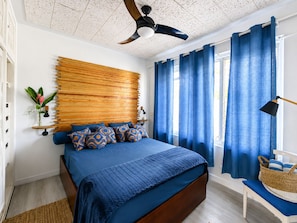 A bedroom with a bed, a ceiling fan, a window with curtains, a bedside table, and a lamp.