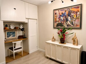Ein Zimmer mit weißem Schreibtisch, einem gerahmten Gemälde, einem Konsolentisch mit Dekoration und einem Teppich.