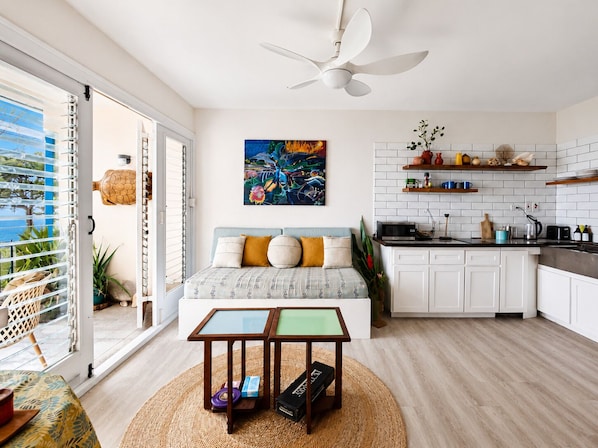 A modern living space with a sofa, a dining table, a kitchen area, and a view of the sea through a sliding door.