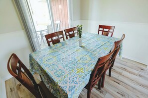 Une table à manger recouverte d’une nappe à motifs, entourée de chaises en bois, dans une pièce dotée d’un plancher en bois et d’une fenêtre avec des stores.