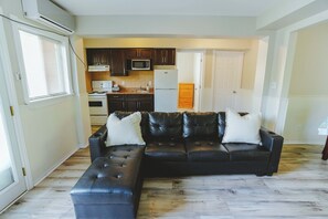 A living room with a black leather sectional sofa, a kitchen with dark wooden cabinets, a white refrigerator, and a microwave. - Heron Cottage - 3-Bedroom Cottage – Family Friendly (Trent Lakes)