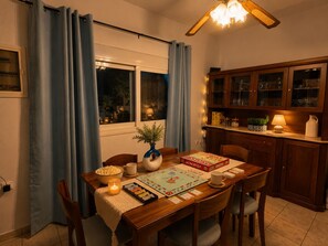 Een eetruimte met een houten tafel, stoelen, een Monopoly spel, een vaas met een plant, en een keuken met kastjes en een lamp.