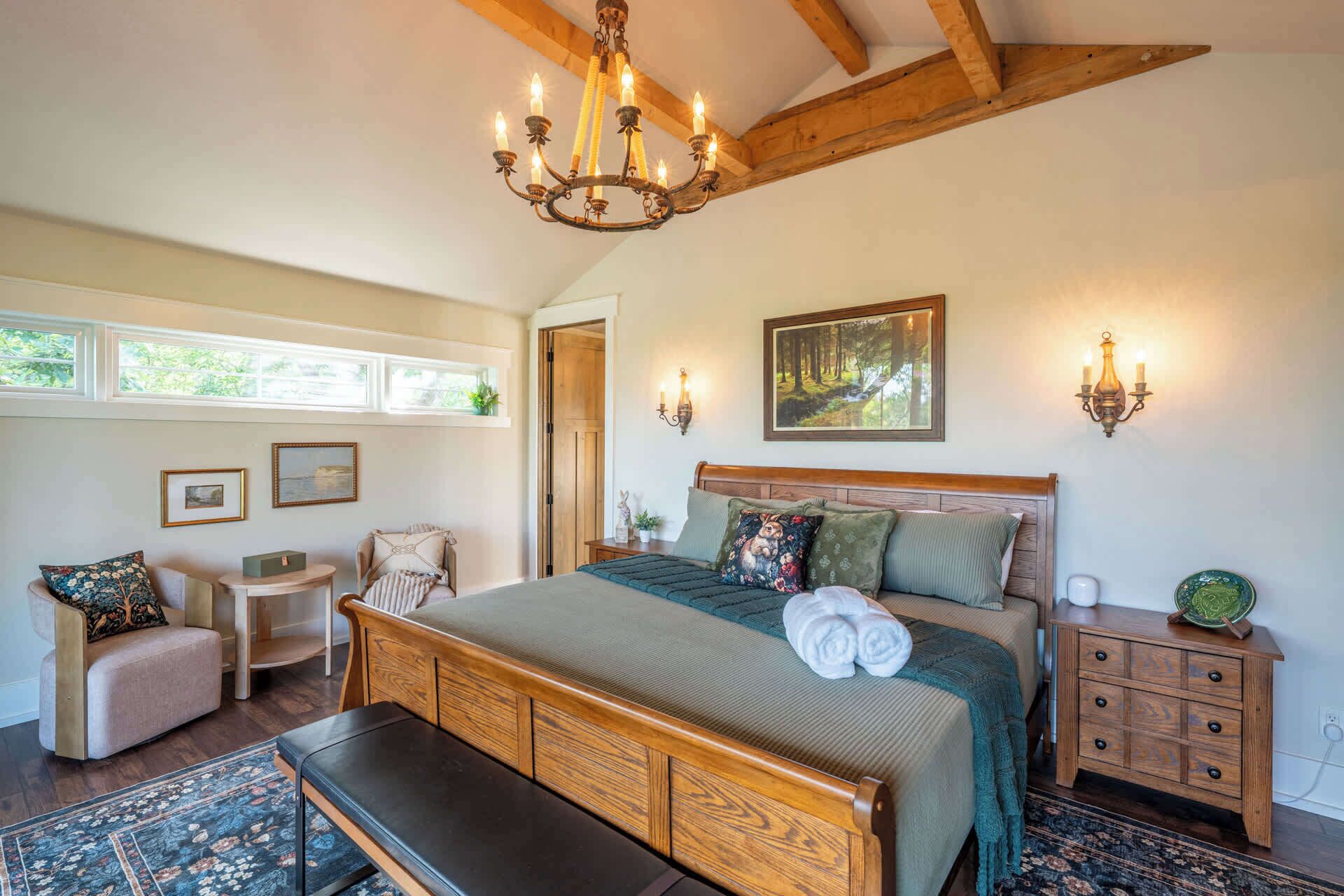 A bedroom with a large bed, a bench, a chair, a nightstand, a wall-mounted lamp, and a painting.