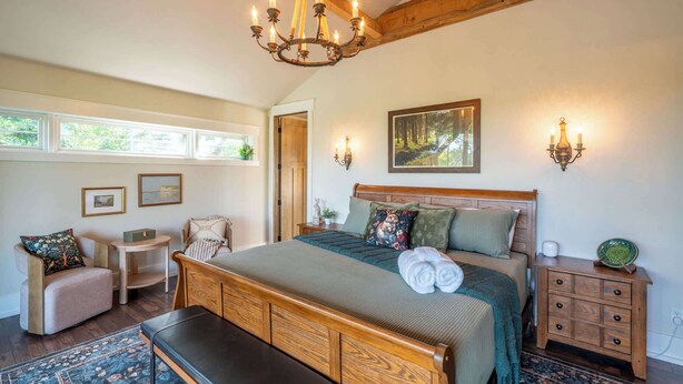 A bedroom with a large bed, a bench, a chair, a nightstand, a wall-mounted lamp, and a painting.