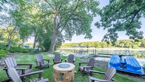 An outdoor seating area with Adirondack chairs and a round wooden table, overlooking a lake with a dock and a blue boat.