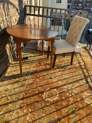 A round wooden table with a cushioned chair on a patterned rug, set against a stone wall and a balcony railing. - Morden King Room in  Boston king (TROPICAL VIBEZ#2) (Boston)