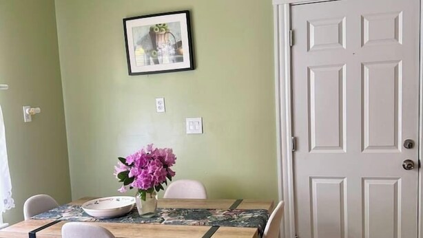 A dining area with a wooden table, four chairs, a vase with flowers, and a framed picture on the wall.