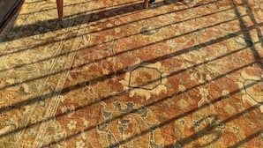 A wooden table and a cushioned chair on a patterned rug, with a metal railing and a building in the background.