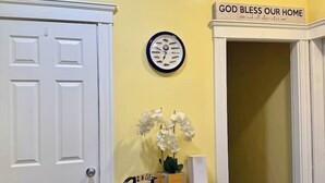 A hallway with a white door, a clock, a small table with a vase of flowers, and a sign above the door that reads 'God bless our home.'