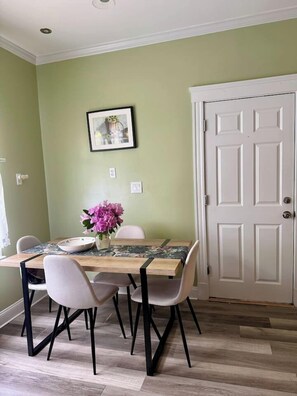 A dining area with a wooden table, four chairs, a vase with flowers, and a framed picture on the wall.