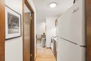 Ein schmaler Flur führt zu einer Küche mit einem Kühlschrank, einer Mülltonne und einem Essbereich mit Tisch und Stühlen.