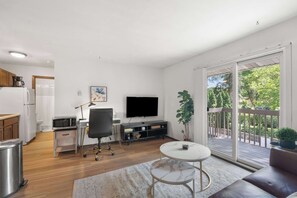 Ett modernt vardagsrum med kök, matplats, en sittgrupp med bord och stolar, en tv och en balkong med utsikt över träd.