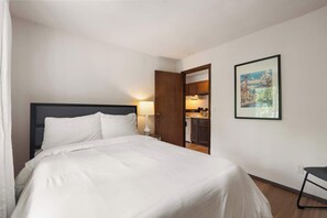 A hotel room with a bed, a nightstand, a lamp, a framed picture on the wall, and an open door to the kitchen. - UW Hospital Retreat with Dedicated Workspace (Madison)