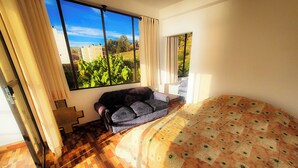 A bedroom with a large bed, a sofa, a window with a view of greenery, and a tiled floor.