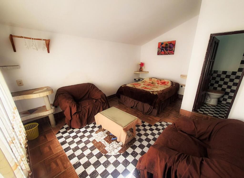 Una habitación pequeña con una cama, una mesita y un sofá. El suelo es de cuadros y hay una estantería con un jarrón.