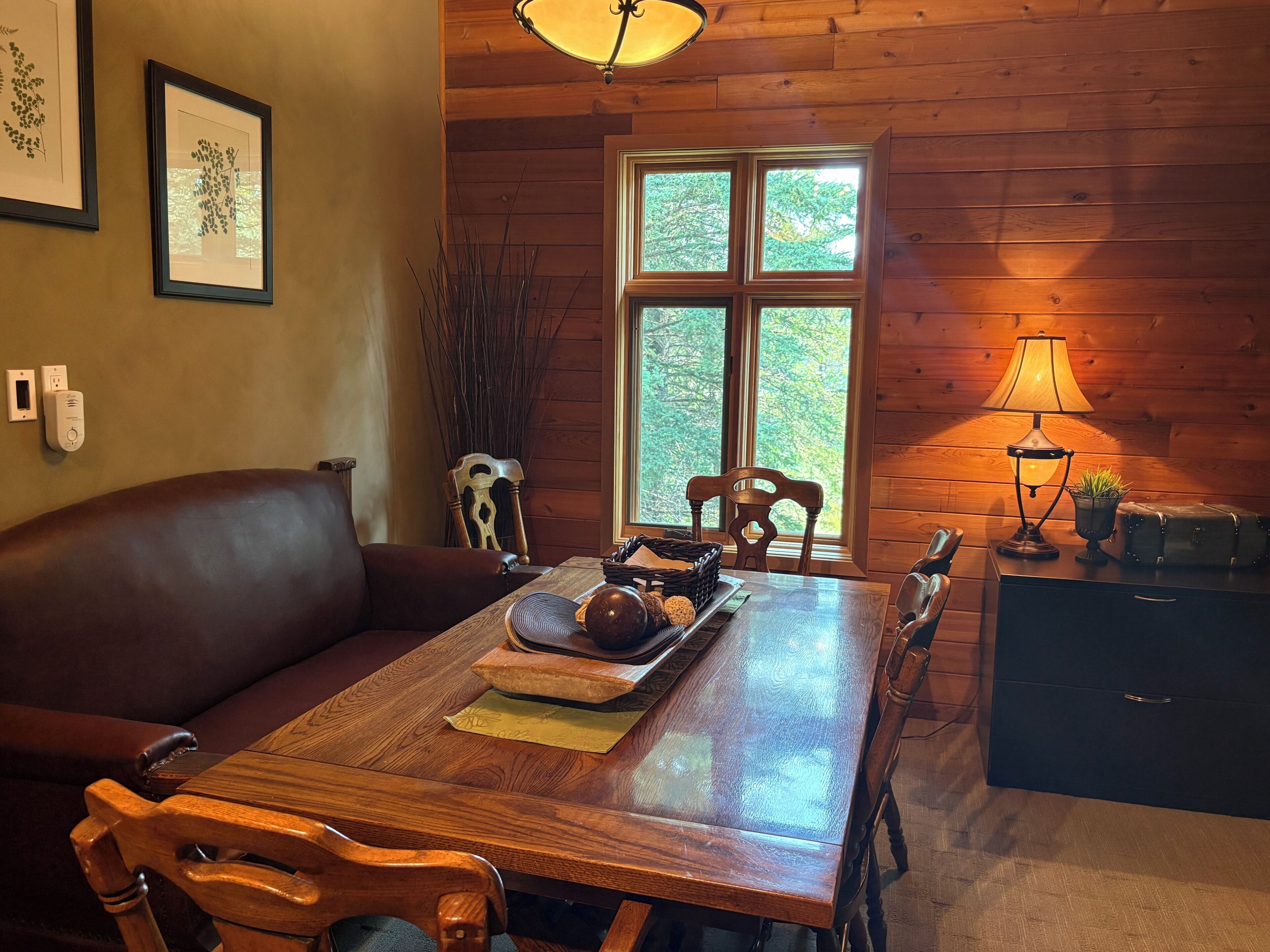 A wooden dining table with chairs, a brown leather couch, a lamp, and a window with a view of trees.