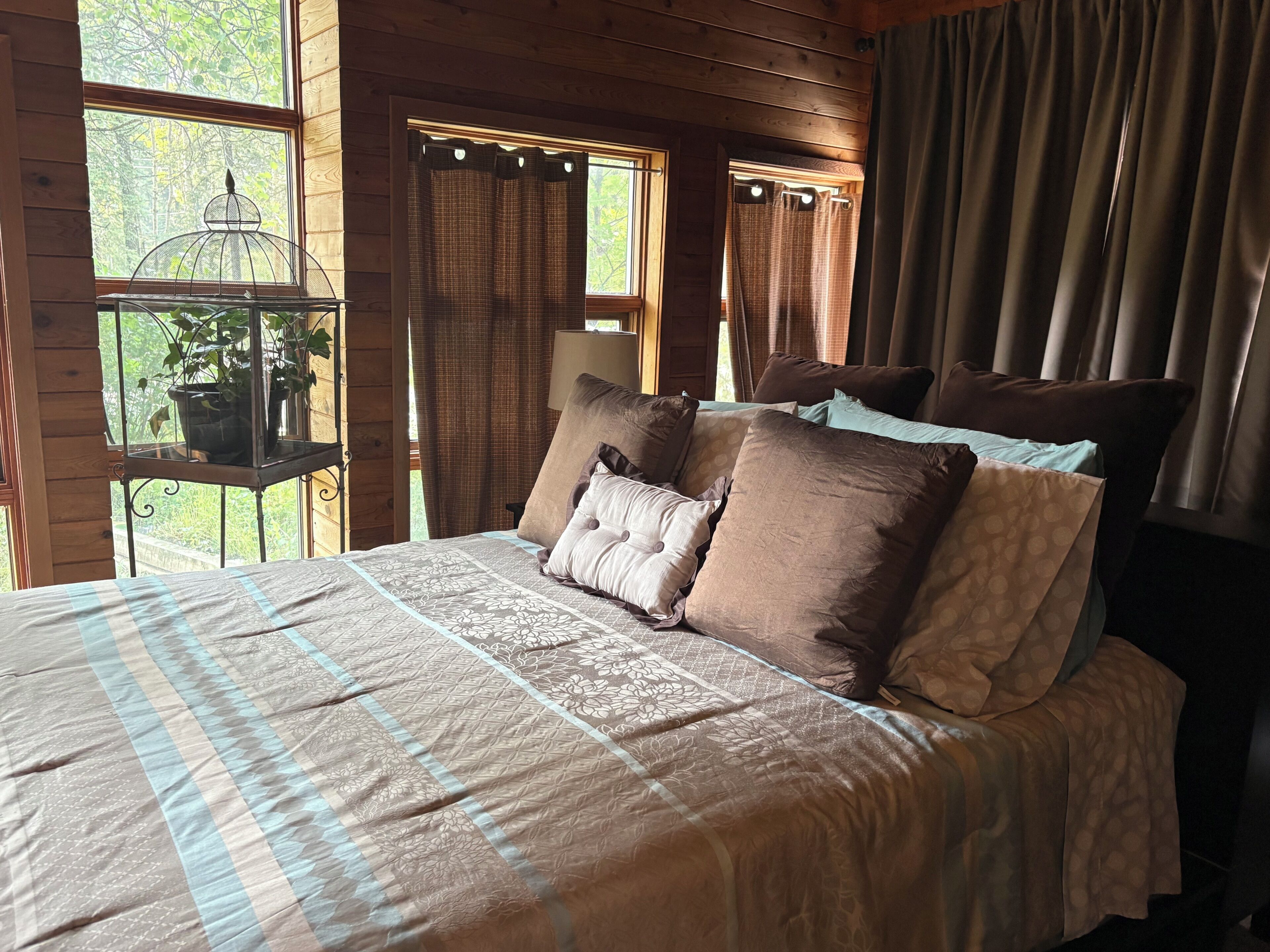 Quarto de cabana de madeira com cama, cortinas e janela com vista para árvores.