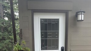 Eine weiße Tür mit einem Metallgitterfenster, ein Holzterrasse und eine graue Wand mit Wandleuchte.