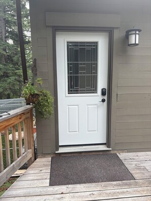 A white door with a metal grid window, a wooden deck, and a gray wall with a wall-mounted light fixture.