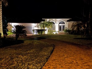 A well-lit residential property at night with a paved driveway leading to a gated entrance, surrounded by landscaped greenery. - Lovely 3-bedroom house with AC in charming Burnt store Marina (Punta Gorda)